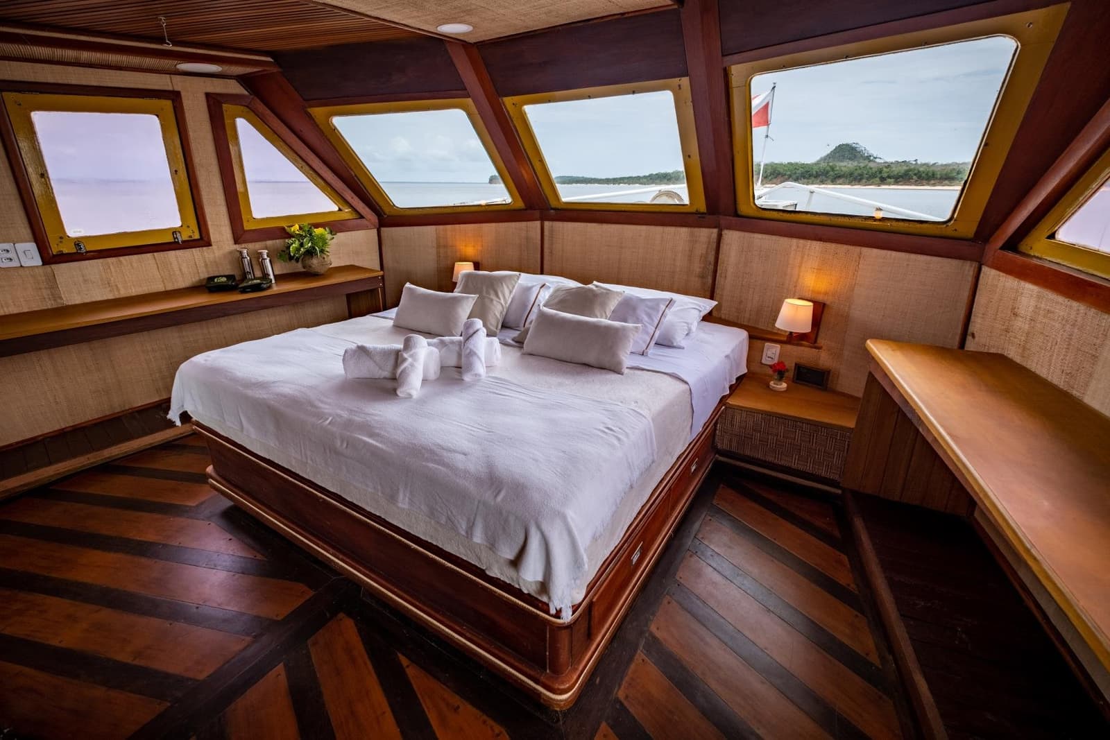 Se réveiller avec le fleuve comme seul horizon, c'est tout l'intérêt. Cabine principale spacieuse à bord d'une croisière Amazonie au départ d'Alter do Chão, avec un grand lit en lin blanc immaculé, des animaux en serviettes pliées, des boiseries chaleureuses et des fenêtres panoramiques donnant sur une rive forestière
