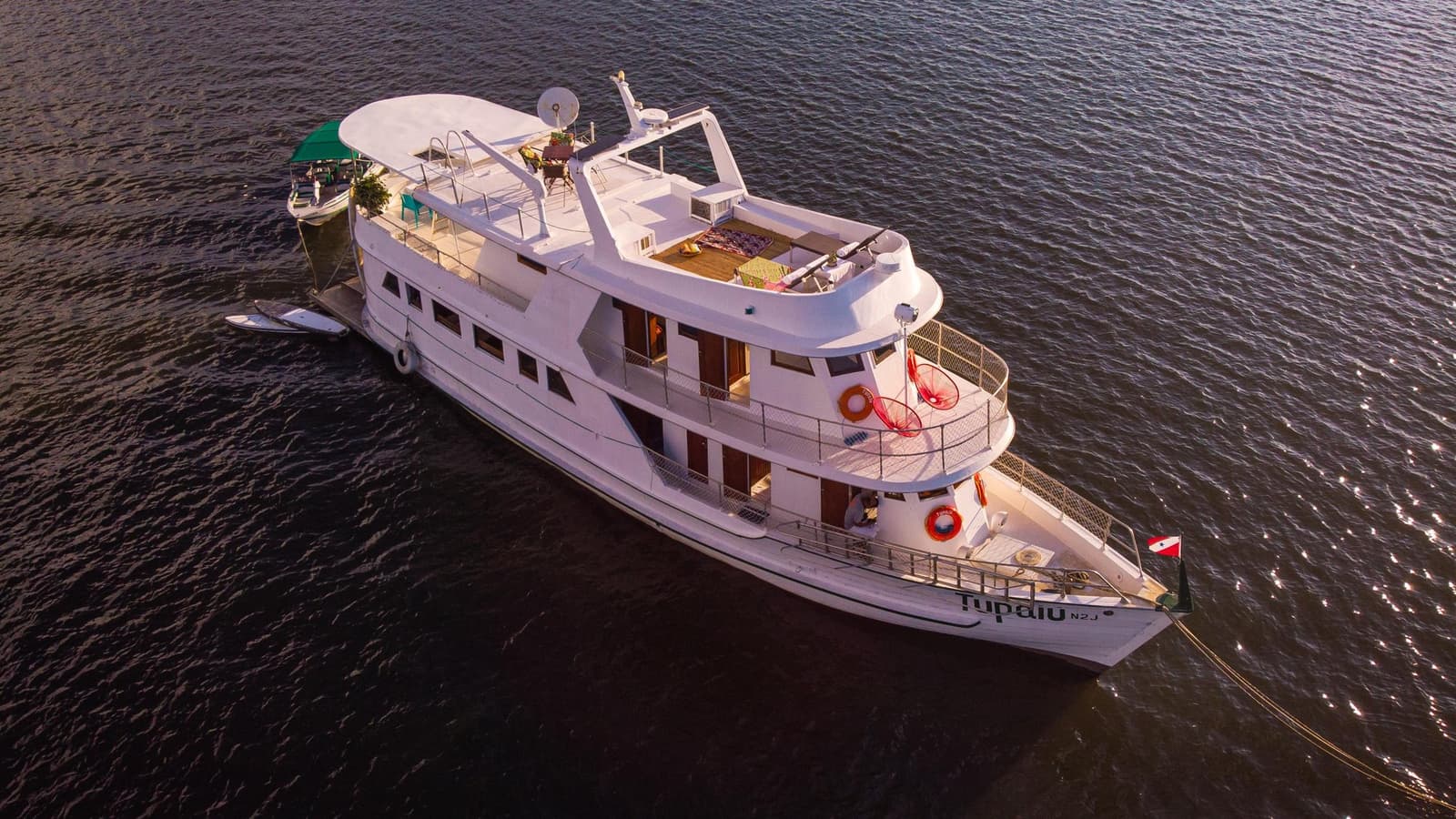 Votre cabine flotte, le fleuve défile, l'aventure commence. Vue aérienne du bateau fluvial blanc à plusieurs ponts Tupaiu sur un large fleuve sombre, avec des ponts soleil ouverts, des espaces détente en teck et une annexe amarrée sur le côté.