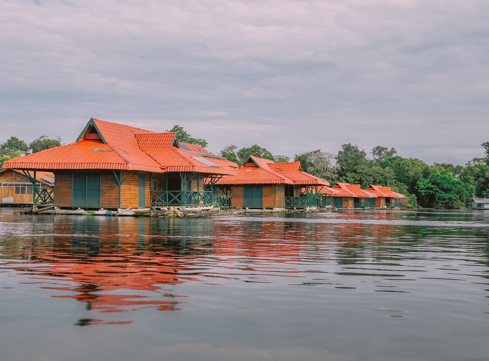 A Amazônia do lado de fora, o rio logo abaixo dos seus pés. Uma fileira de cabanas de madeira sobre a água com telhados de telha laranja refletidos nas águas calmas do rio, cercadas pela densa floresta tropical amazônica