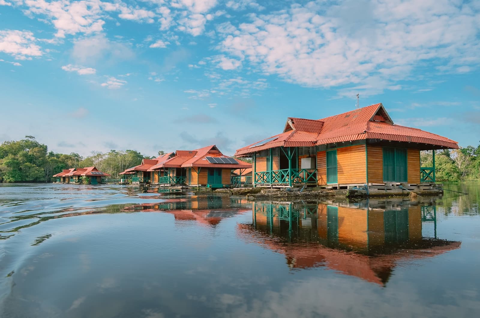 A varanda dá pro rio. O rio dá pra floresta. A floresta dá pra tudo. Uma fileira de cabanas flutuantes de madeira com energia solar, telhados vermelhos e grades teal refletidas na água escura e calma de uma viagem de ecolodge na floresta amazônica no Brasil, cercada por densa vegetação ribeirinha sob céu azul