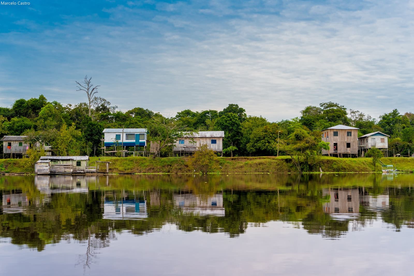 Palafitas, silêncio e o rio fazendo o espelho, a Amazônia no seu ritmo. Casas de madeira sobre palafitas refletidas em um canal de rio calmo, com densa cobertura de floresta tropical ao fundo — uma comunidade ribeirinha visitada durante uma viagem de ecolodge na floresta amazônica no Brasil