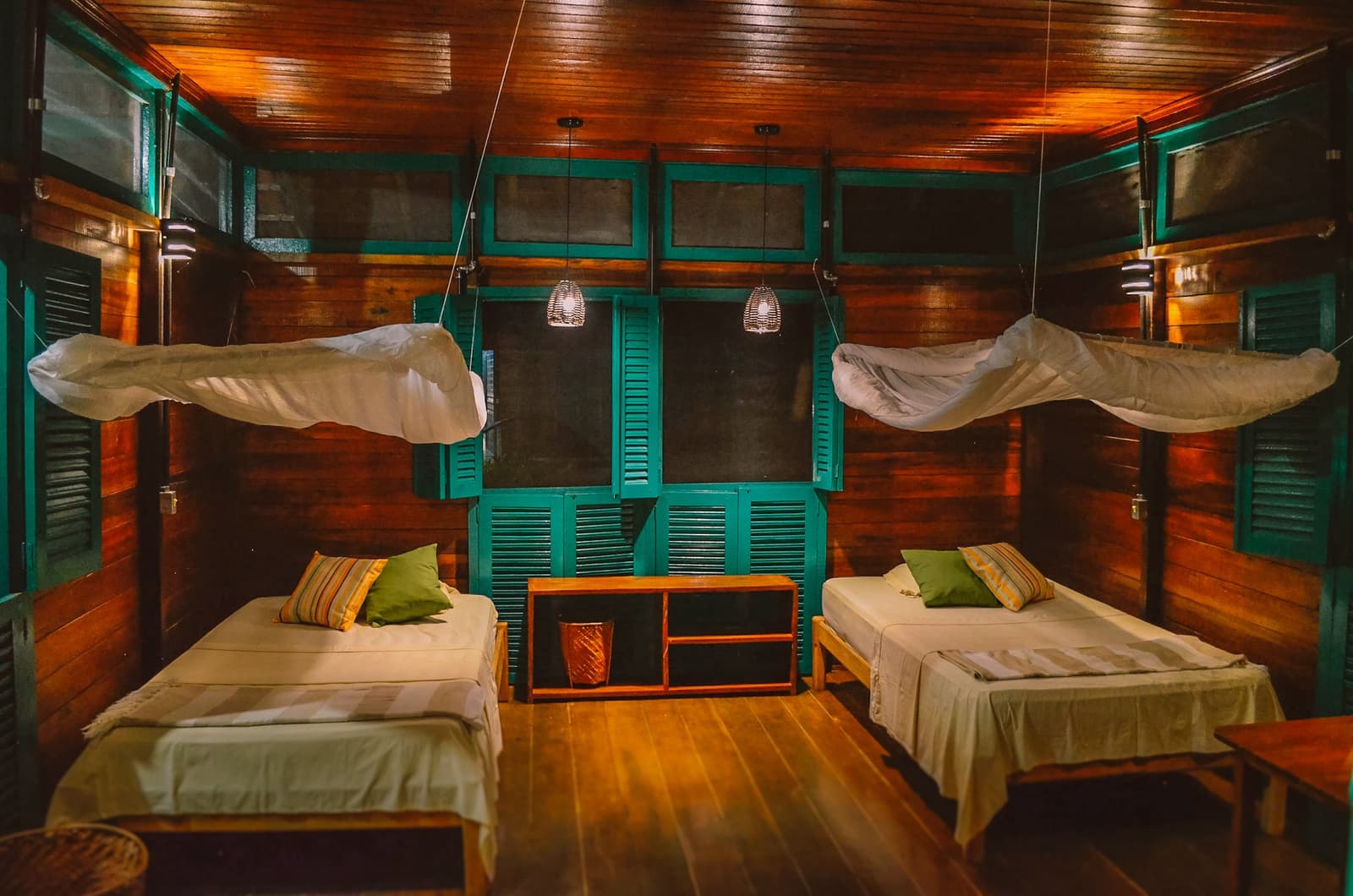 Madeira de lei, venezianas verdes e mosquiteiro: a Amazônia tem seu jeito de receber bem. Quarto duplo em um ecolodge na floresta amazônica no Brasil com mosquiteiros suspensos, paredes de madeira aquecida e janelas com venezianas turquesas