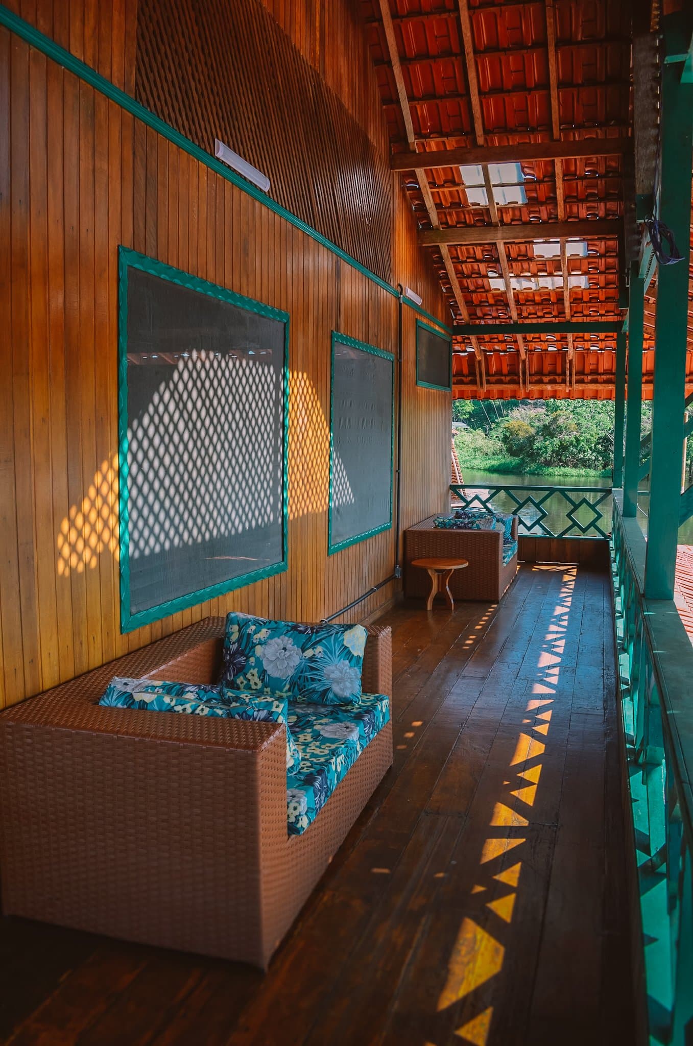 Rio à vista, brisa constante e o sofá perfeito para não fazer nada. Varanda de madeira coberta de um ecolodge na floresta amazônica no Brasil, com poltronas de vime e almofadas de estampa tropical, luz filtrada em xadrez sobre o assoalho e uma paisagem fluvial verde ao fundo do corredor