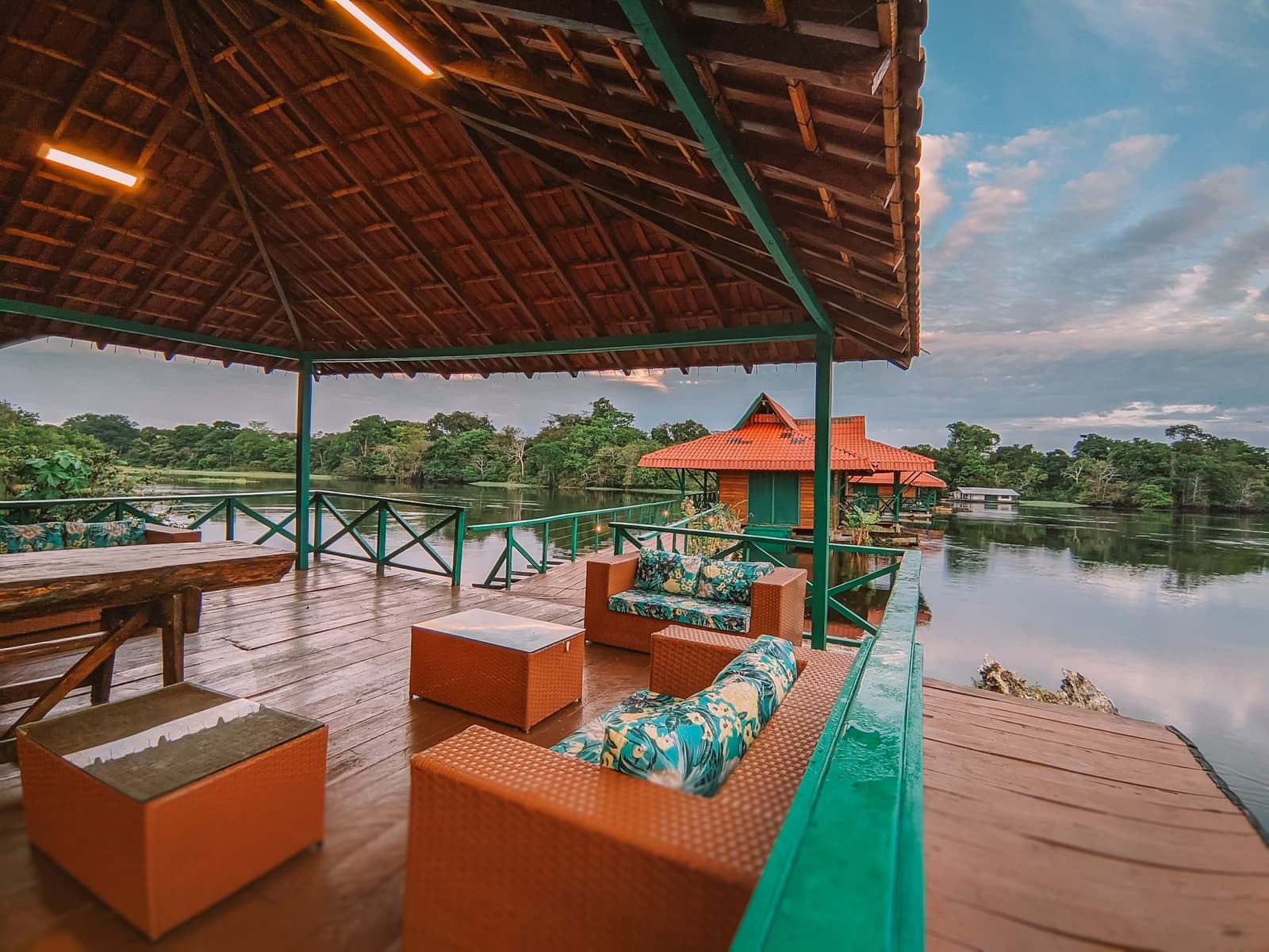 Aquele lugar onde o entardecer chega antes de você perceber. Terraço coberto de madeira com móveis de vime voltado para um rio calmo e densa floresta em um passeio de ecolodge na floresta amazônica no Brasil