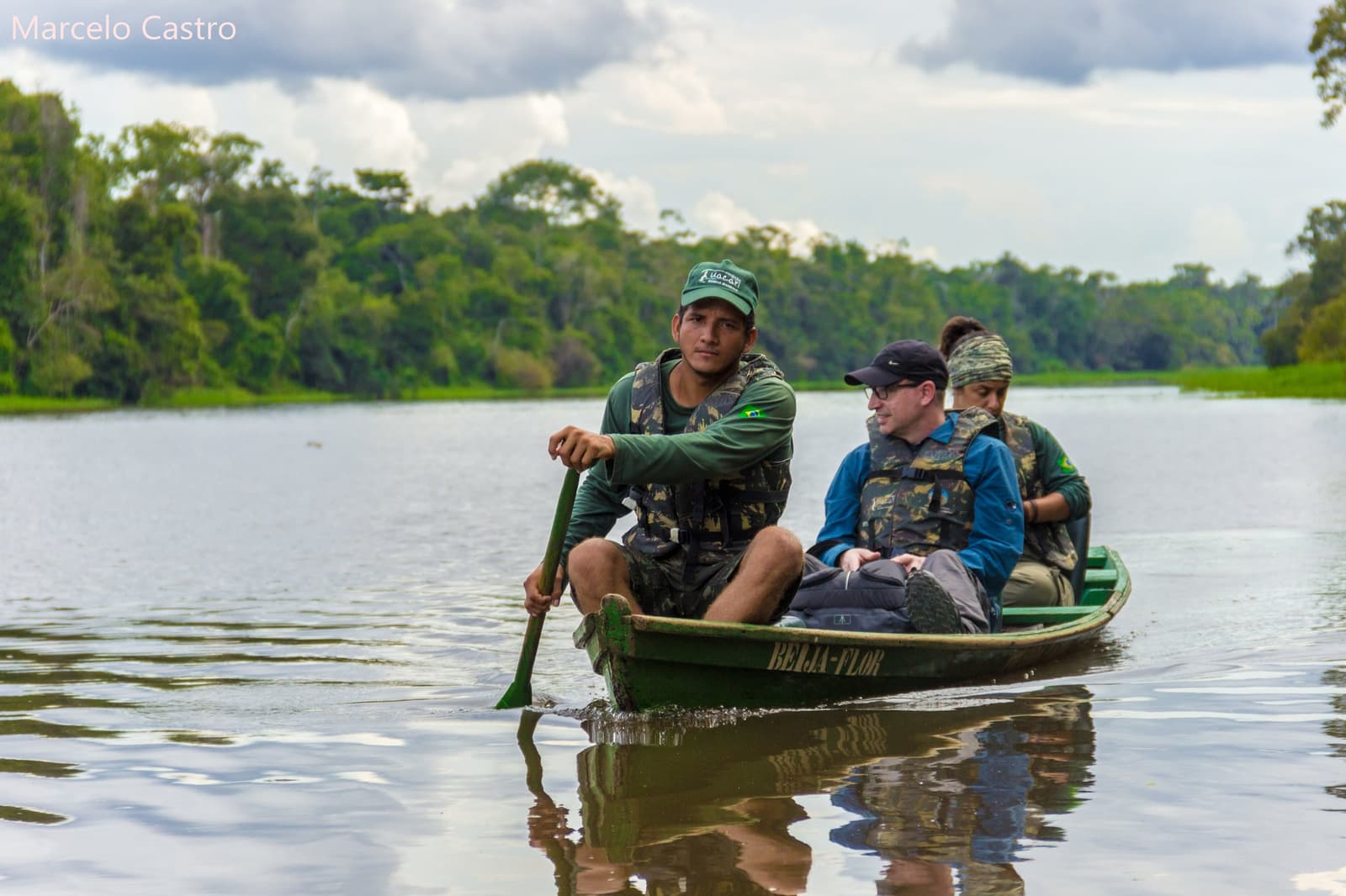 Rio calmo, remo firme, e a floresta ouvindo tudo. Um guia local rema uma canoa de madeira chamada Beija-Flor com dois ecoturistas por um canal fluvial calmo, com densa floresta de várzea ao fundo