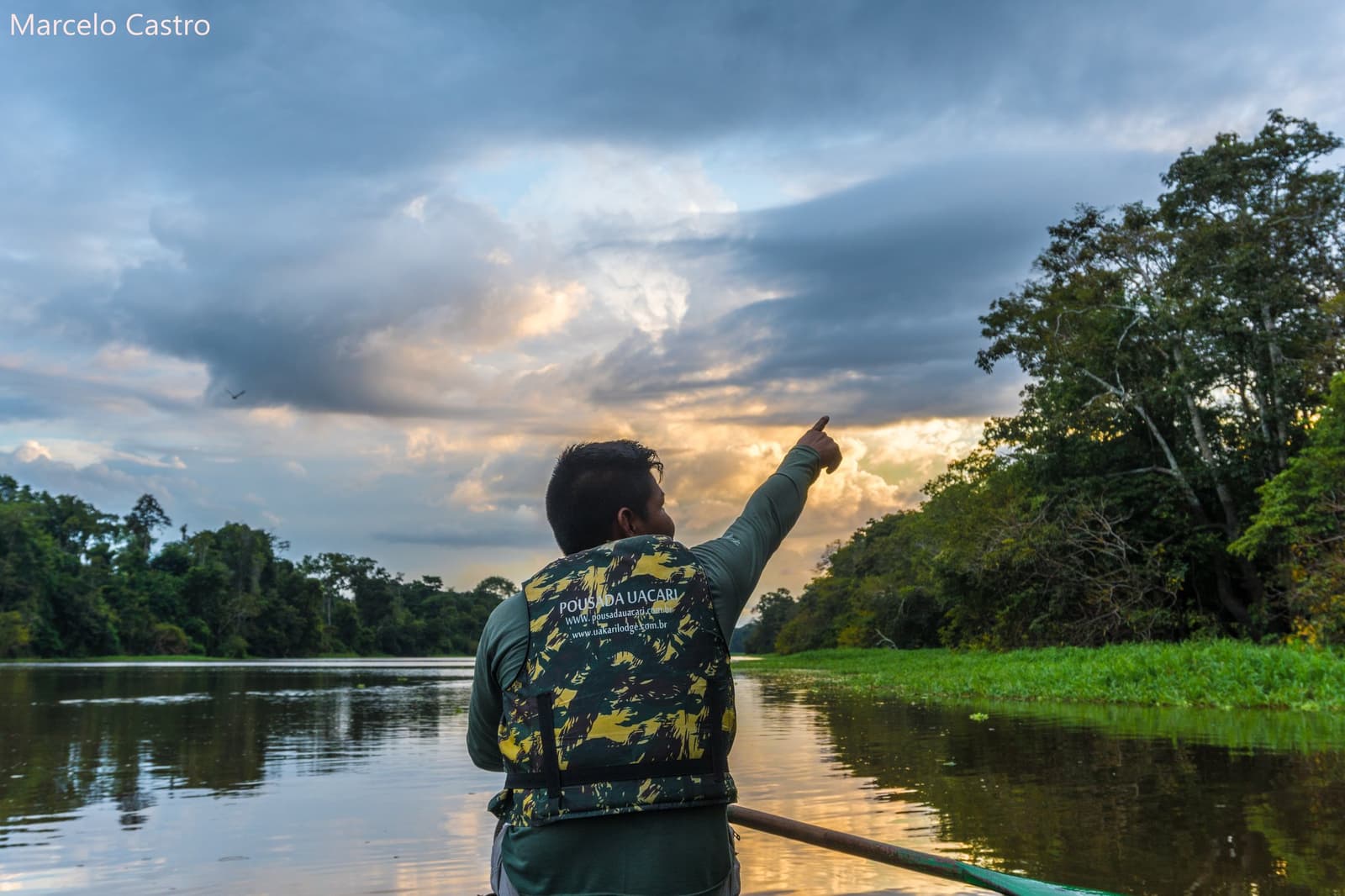 Ele aponta, você olha, e de repente tudo faz sentido. Um guia da Pousada Uacari aponta para o horizonte iluminado a partir de um barco em um canal tranquilo da Amazônia, cercado por floresta densa ao pôr do sol
