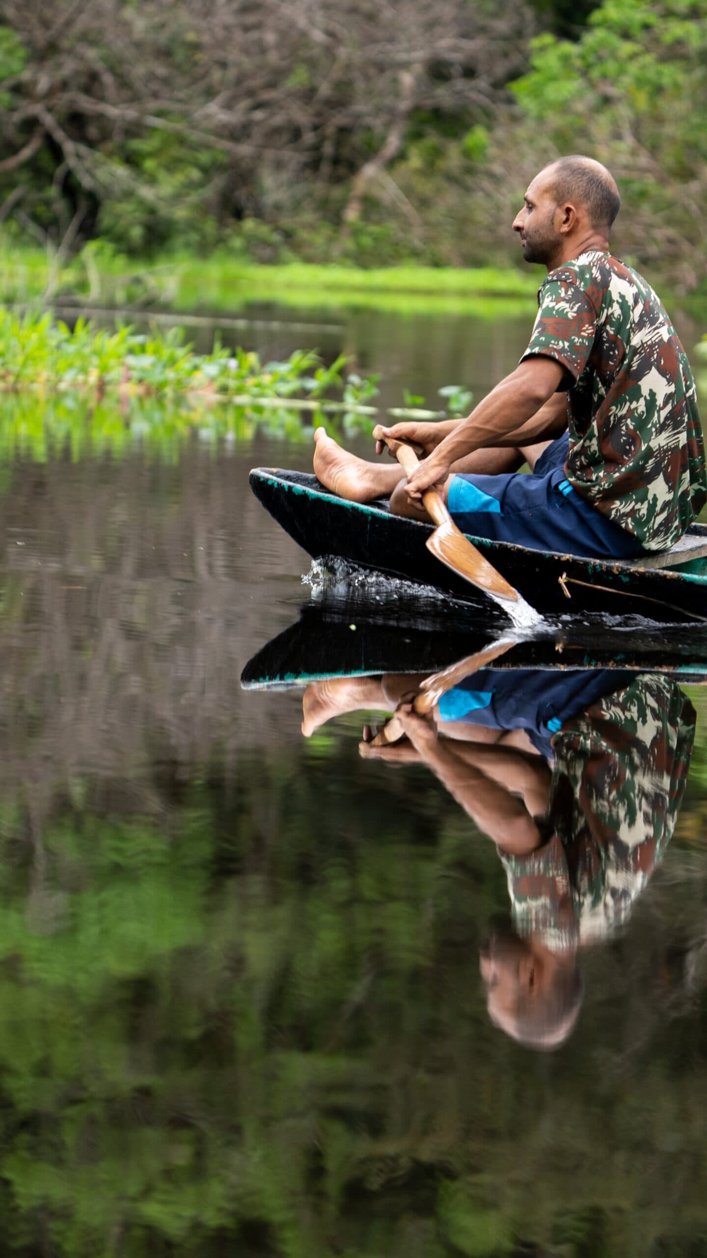 Cada remada conta uma história que nenhum roteiro consegue traduzir. Um homem rema uma canoa de madeira tradicional sobre águas completamente calmas durante um tour de sobrevivência na Amazônia, seu reflexo perfeitamente espelhado na superfície escura ao lado de vegetação ribeirinha exuberante