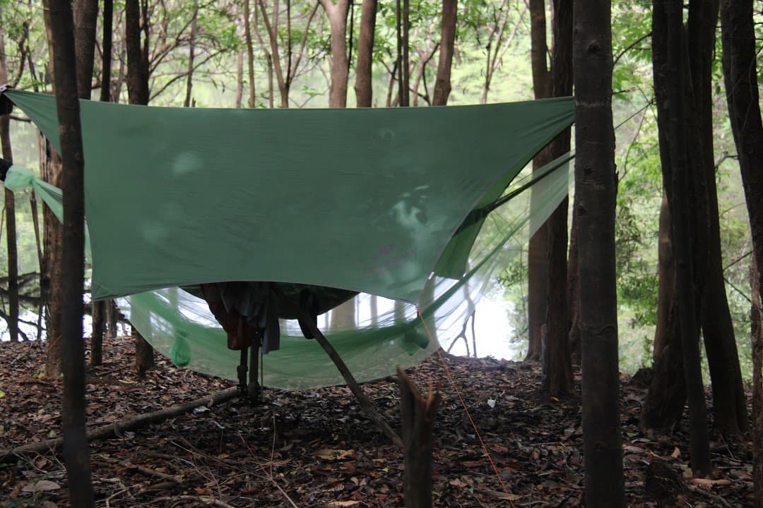 Quarto na mata: a floresta cuida do ar condicionado. Lona verde armada entre árvores sobre uma rede com mosquiteiro em um tour de sobrevivência na Amazônia, com folhiço no chão e água visível ao fundo entre os troncos