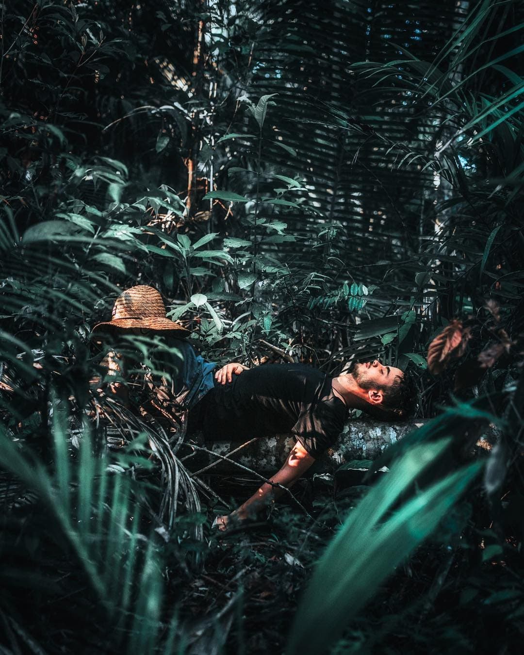 A floresta pede silêncio, e o corpo logo concorda. Um homem recostado sobre um tronco coberto de musgo no meio de uma densa vegetação de floresta tropical durante um tour de sobrevivência na Amazônia, com seu chapéu de palha ao lado entre folhas tropicais