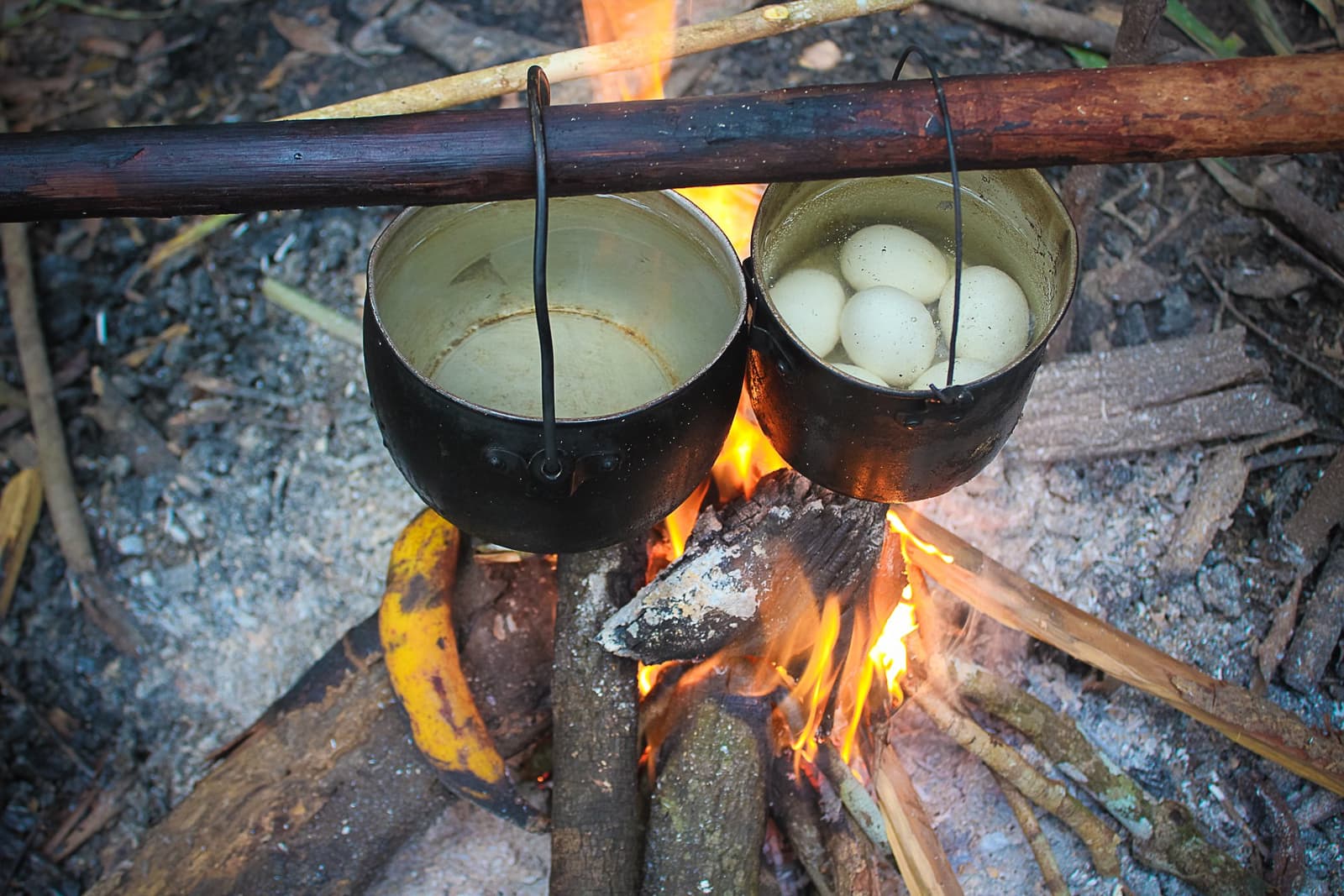 Kochen auf die einfache Art, Feuer, Topf, fertig. Zwei geschwärzte Töpfe hängen über einem offenen Lagerfeuer im Dschungel, einer mit kochenden Eiern