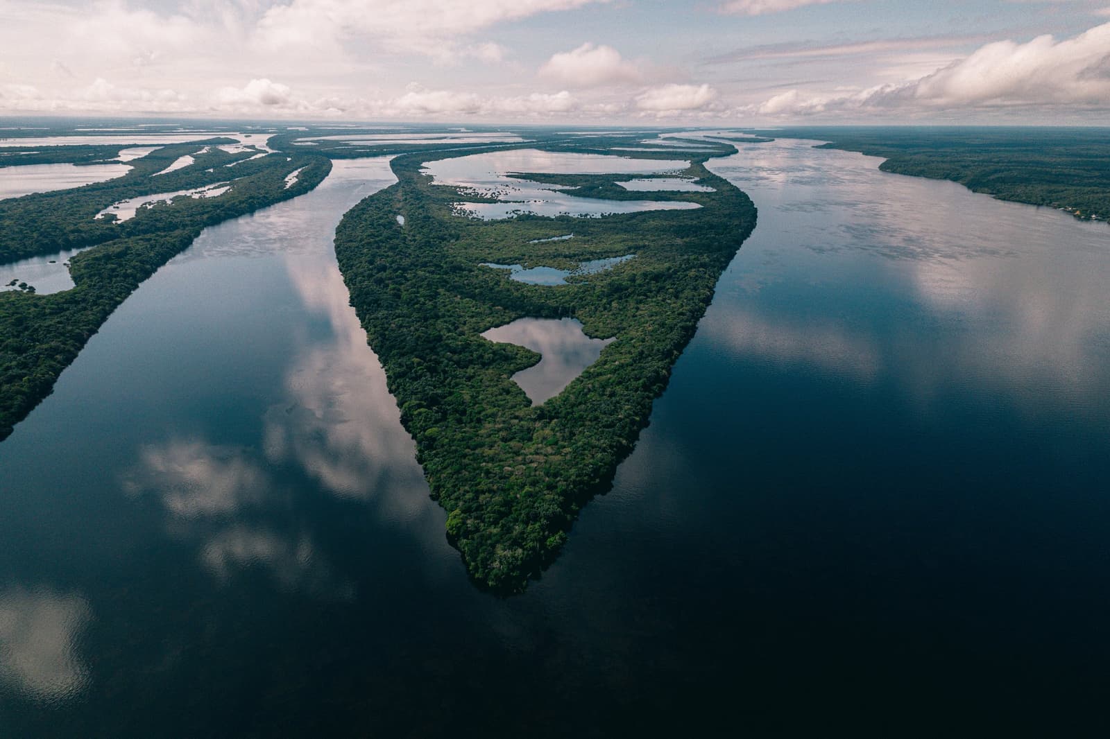 Rio, ilha, céu, reflexo, tudo junto e misturado, do jeito que a Amazônia gosta. Vista aérea de uma ilha densamente florestada no Anavilhanas Jungle Lodge Brasil, rodeada pelas águas escuras e calmas de um vasto arquipélago fluvial sob um céu nublado