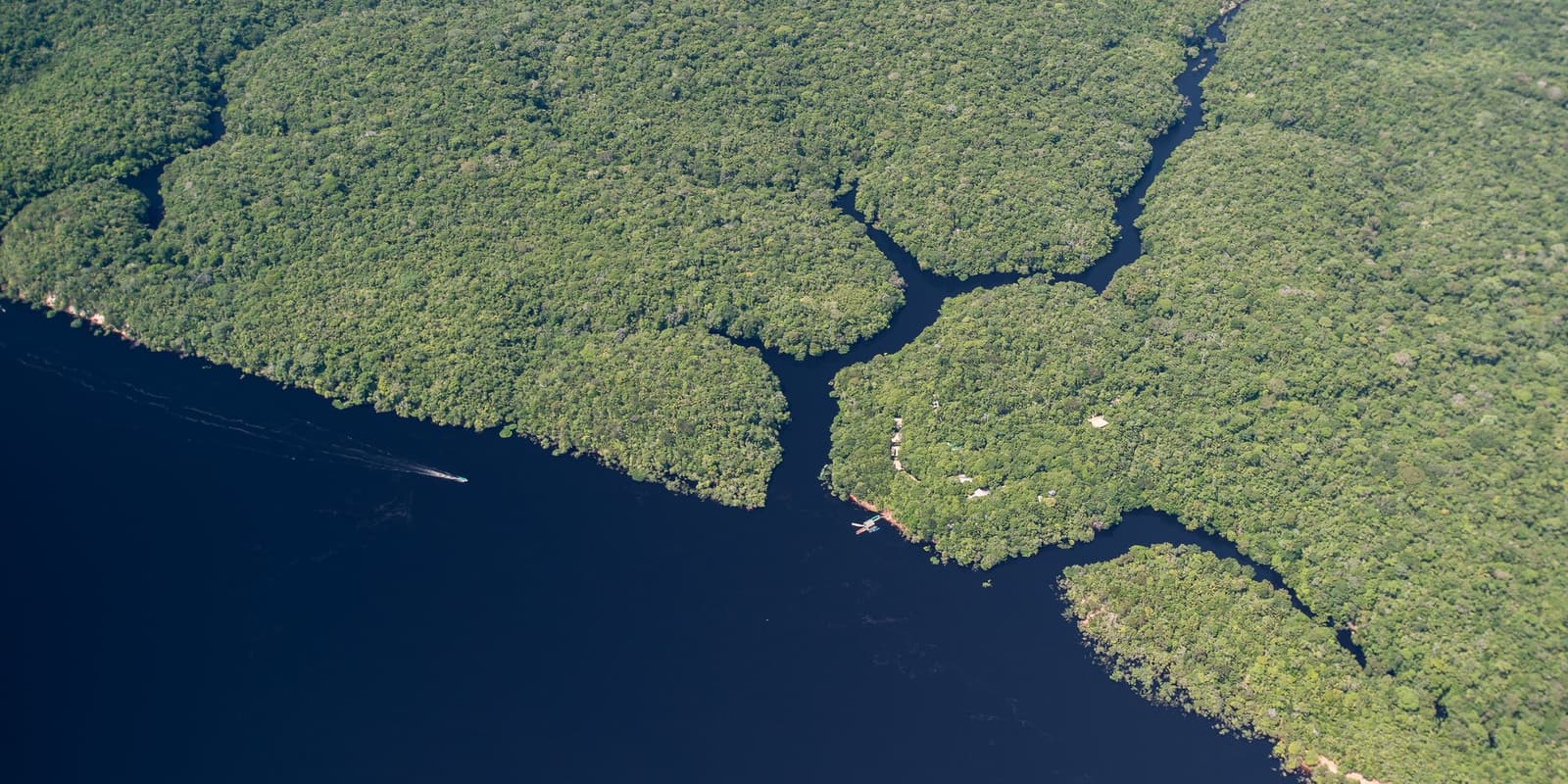 O rio não separa a floresta, ele faz parte dela. Vista aérea do Anavilhanas Jungle Lodge Brazil à beira das águas escuras do Rio Negro, cercado por uma copa densa e ininterrupta da floresta amazônica com estreitos canais serpenteando entre ilhas cobertas de vegetação, um pequeno barco deixando rastro na água