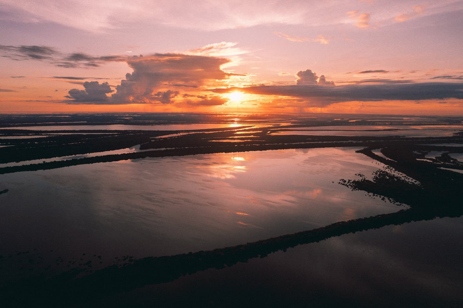 O rio guarda cada cor que o céu resolve soltar. Vista aérea de uma vasta várzea amazônica inundada ao pôr do sol, com luz laranja e rosa refletida sobre igarapés entrelaçados e ilhas florestais escuras, próximo ao Anavilhanas Jungle Lodge Brasil