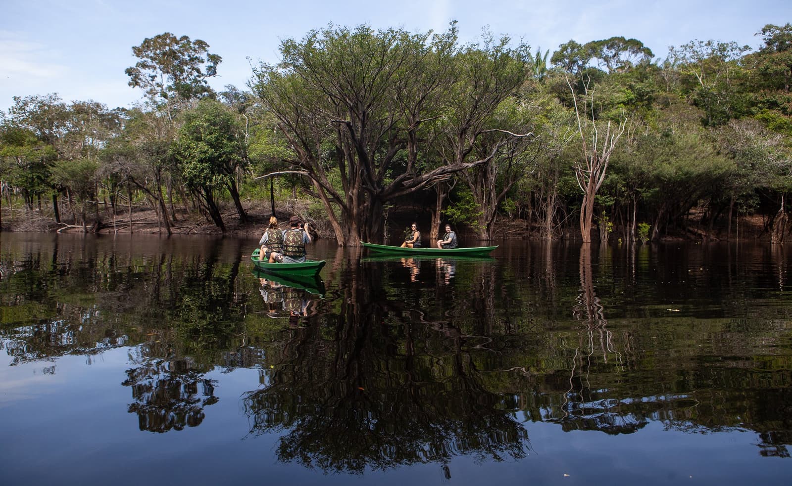 Dentro do igapó, a canoa some devagar entre os galhos. Três viajantes em uma canoa verde deslizando por uma floresta alagada em águas escuras e calmas na Anavilhanas Jungle Lodge Brasil, cercados por árvores parcialmente submersas e seus reflexos perfeitos