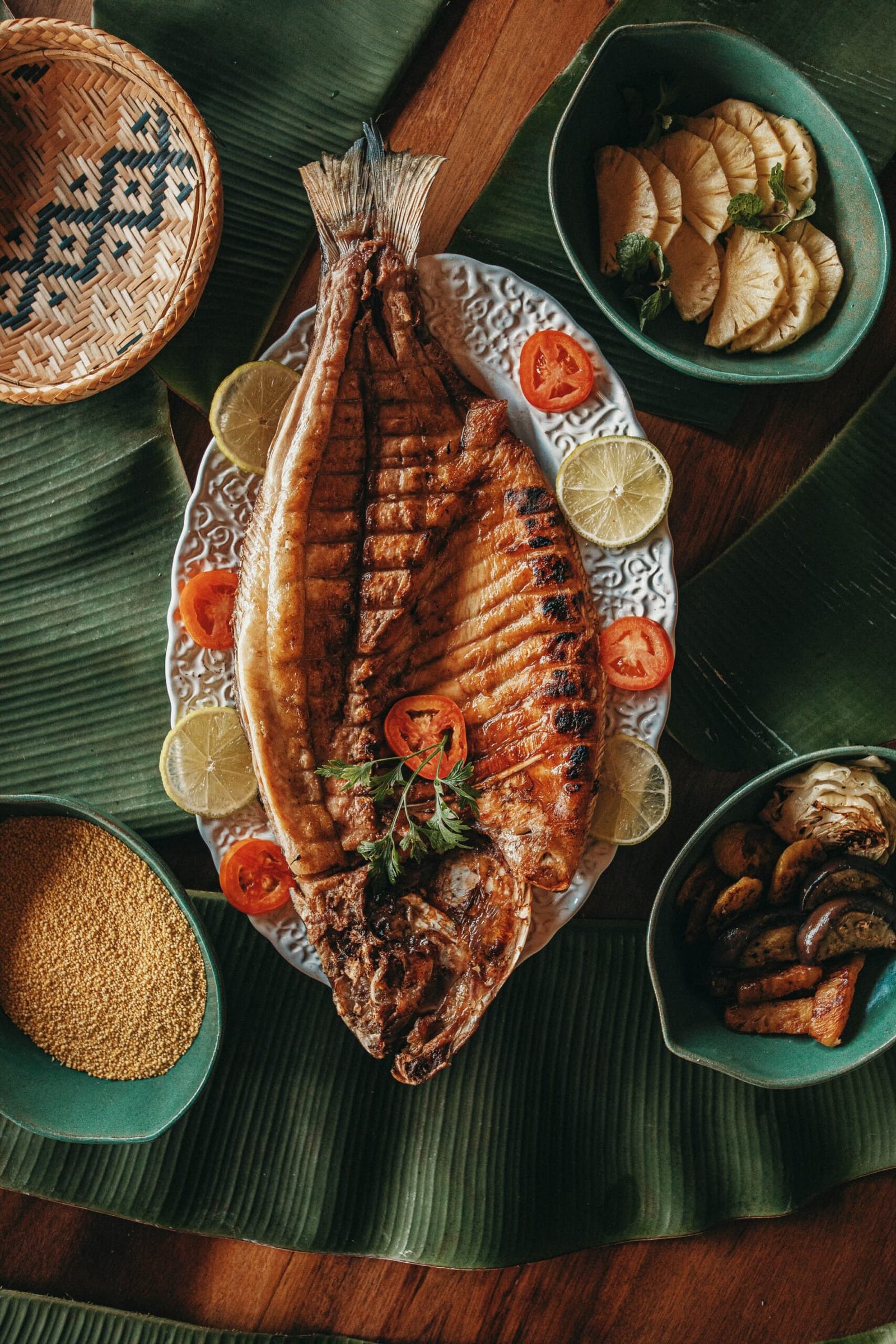Peixe do rio, farofa na mesa: a Amazônia no prato. Um peixe inteiro grelhado servido em prato branco decorativo com rodelas de limão e tomate na Anavilhanas Jungle Lodge Brasil, cercado por tigelas de abacaxi, cogumelos assados e sementes douradas sobre folhas de bananeira