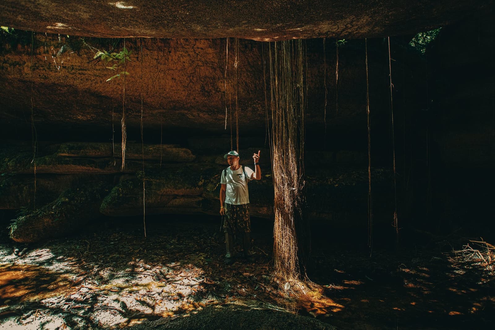 Debaixo da pedra, a floresta ainda encontra um jeito de crescer. Um guia aponta para cima sob uma imensa rocha antiga nas Cavernas do Madadá, Anavilhanas Jungle Lodge Brasil, com longas raízes aéreas caindo do teto em uma dramática luz dourada