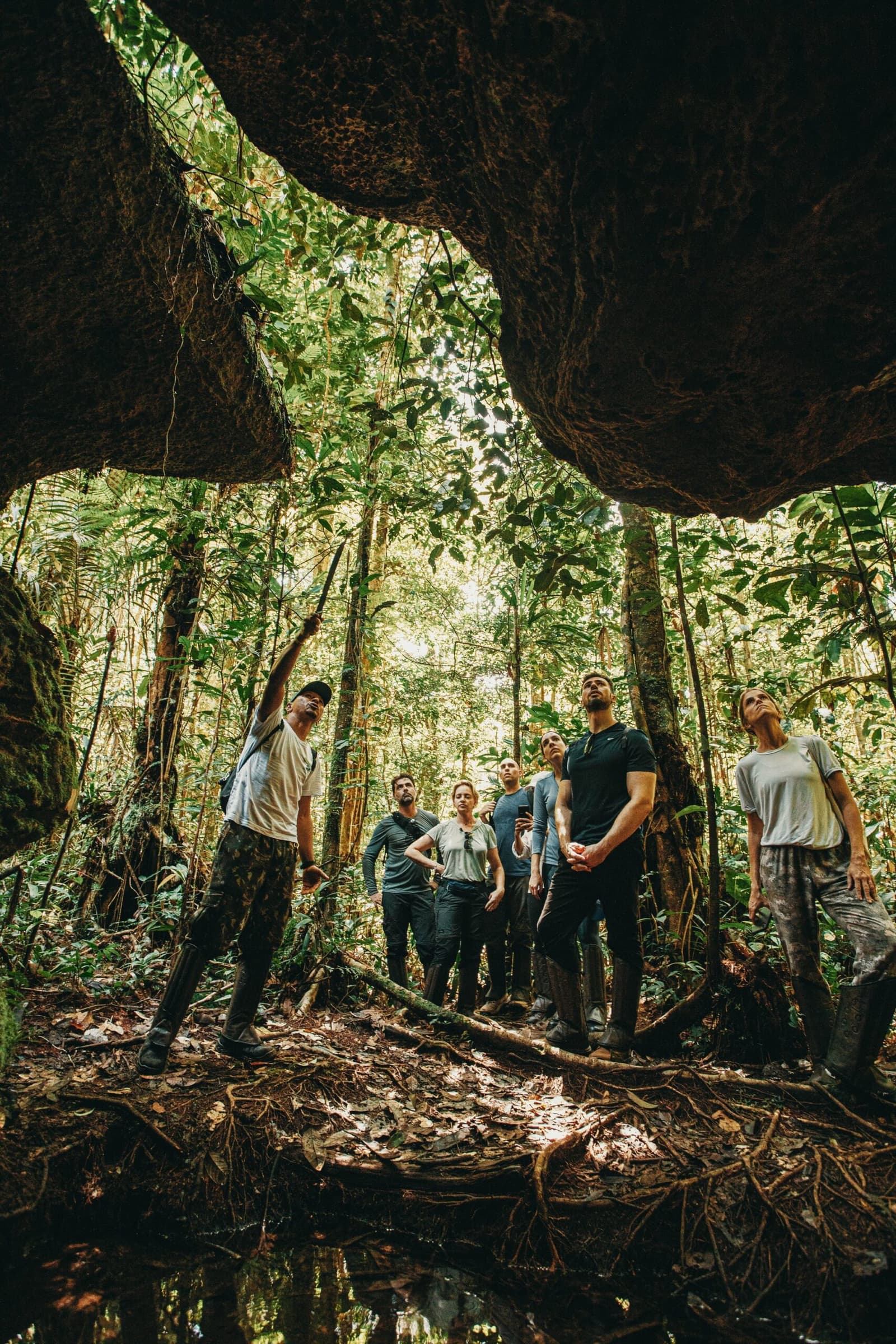 A floresta não precisa de legenda, ela fala por conta própria. Um guia aponta para cima enquanto um pequeno grupo de viajantes contempla a copa da floresta tropical emoldurada pelo arco rochoso de uma caverna no Anavilhanas Jungle Lodge Brasil