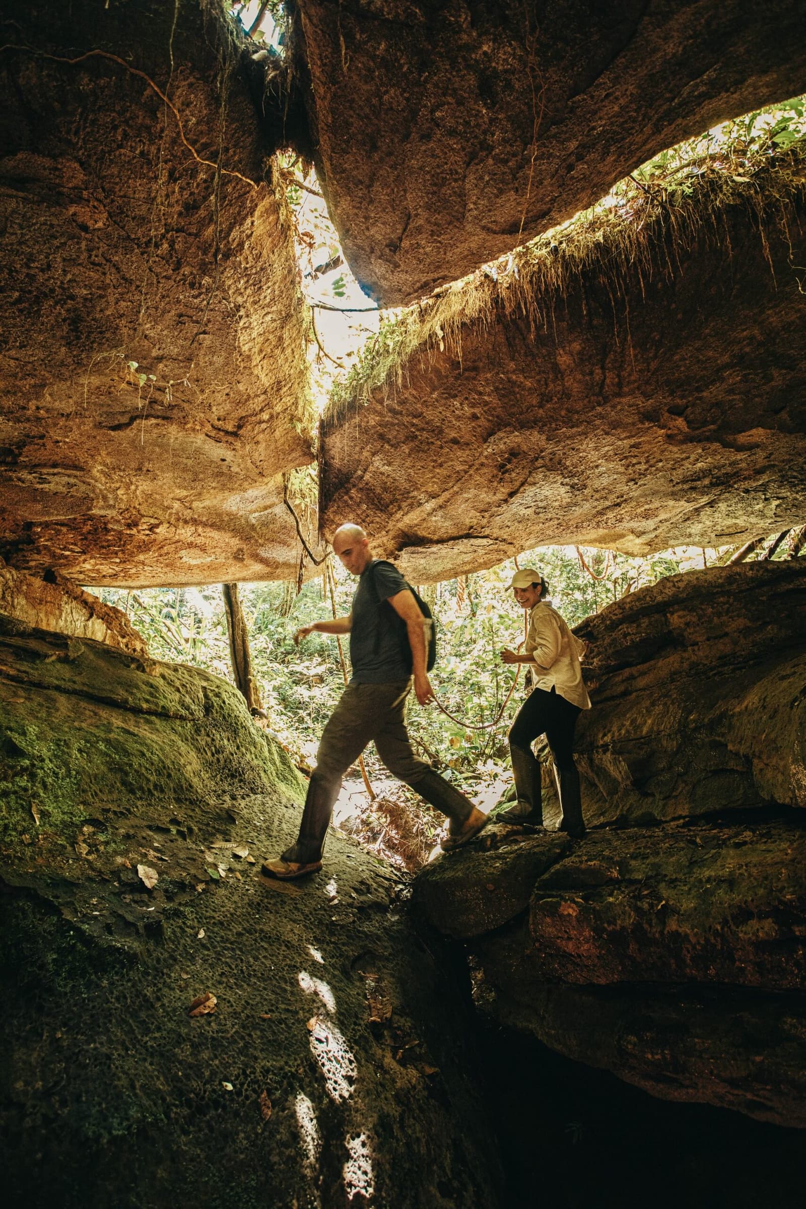 Tem trilha que pede atenção em cada passo, e vale cada um deles. Dois caminhantes se curvam sob uma enorme saliência de arenito nas Cavernas do Madadá durante uma excursão do Anavilhanas Jungle Lodge Brazil, com a selva iluminada visível pela abertura na rocha