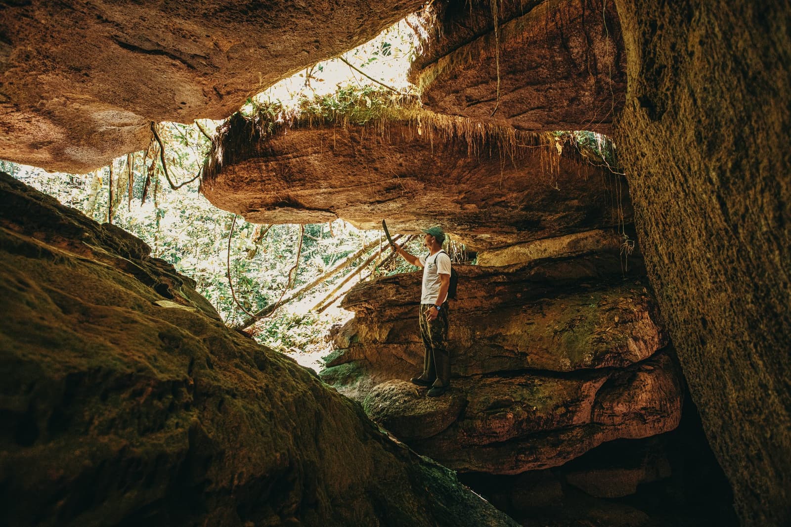 A floresta esconde salas que nenhum arquiteto conseguiria imaginar. Um guia está de pé no interior das antigas formações rochosas das Cavernas do Madadá durante uma trilha a partir do Anavilhanas Jungle Lodge Brazil, com o olhar voltado para o teto de rocha em balanço coberto de raízes suspensas