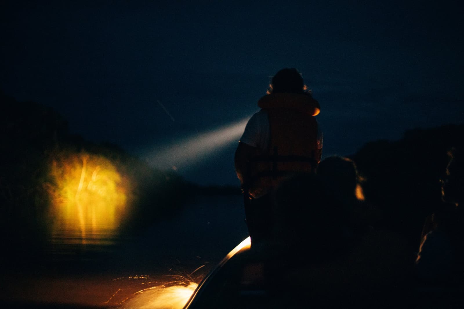 A noite na Amazônia não é silêncio, é espera. O farol faz a pergunta. Um guia em silhueta com colete salva-vidas varre um holofote sobre as águas escuras do rio durante um passeio noturno no Anavilhanas Jungle Lodge Brasil, com uma luz amarela quente brilhando na proa