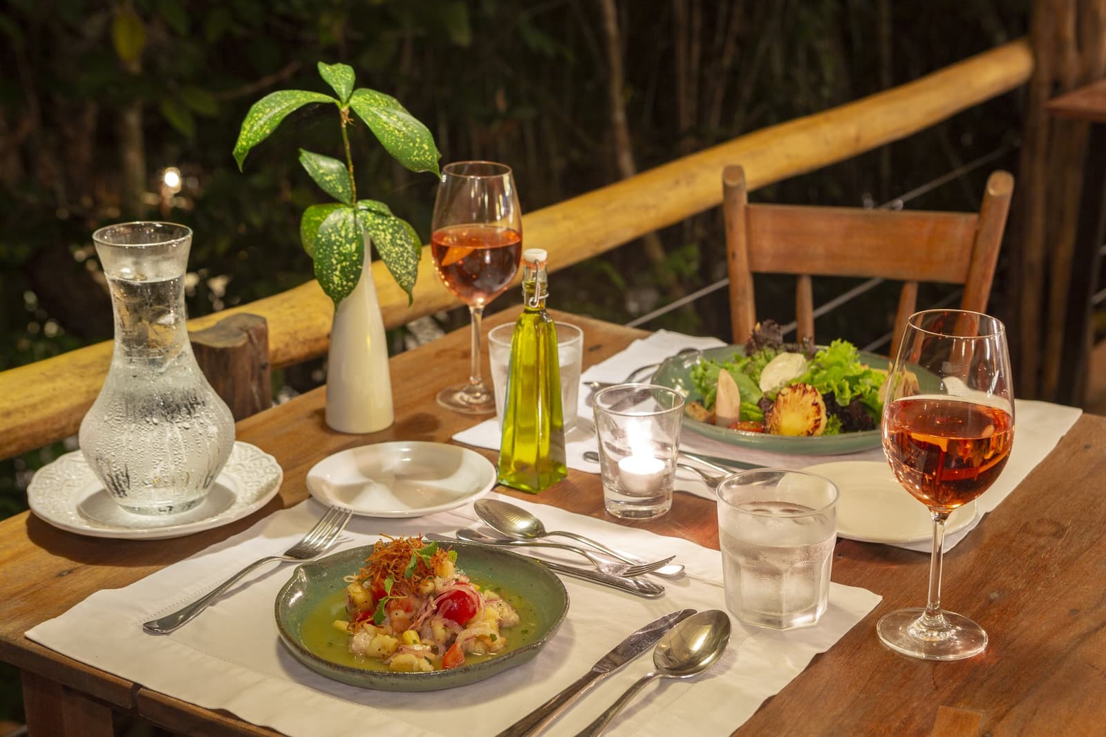 Comida feita com cuidado, vinho gelado, e a floresta como pano de fundo. Mesa de jantar ao ar livre à luz de velas no Anavilhanas Jungle Lodge Brazil, com vinho rosé, um prato de cerâmica verde com entrada de frutos do mar, salada fresca, azeite e uma folha tropical como centro de mesa em um terraço com corrimão de bambu