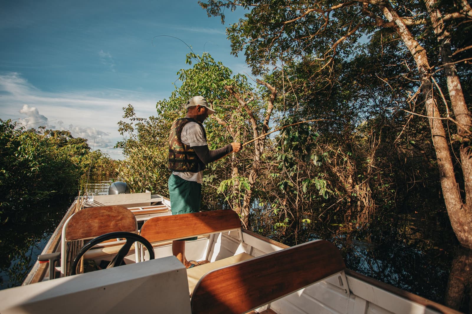 Vara de bambu, água preta e um silêncio que o rio impõe. Um guia em pé na proa de um pequeno barco a motor, com vara de pesca na mão, lançando a linha em direção à vegetação ribeirinha densa durante uma pescaria recreativa no Anavilhanas Jungle Lodge Brasil