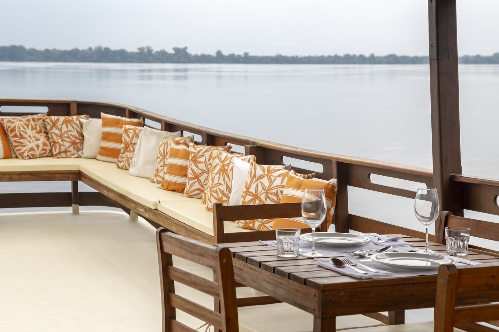 Almoçar com o rio na frente e a floresta ao fundo: difícil de superar. Mesa posta com taças de vinho e talheres no deck aberto de um barco no Anavilhanas Jungle Lodge Brazil, com um sofá lounge almofadado e um rio largo se estendendo até o horizonte coberto de floresta