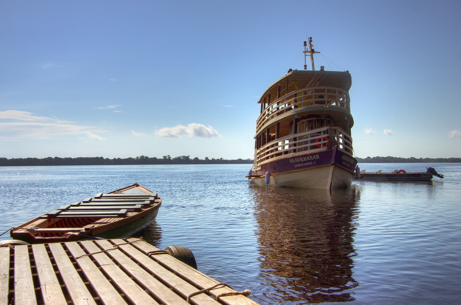 O barco pronto, o rio quieto. Só falta você embarcar. O barco da Anavilhanas Jungle Lodge Brasil ancorado nas águas negras e calmas do Rio Negro, visto de um trapiche de madeira com uma canoa tradicional em primeiro plano