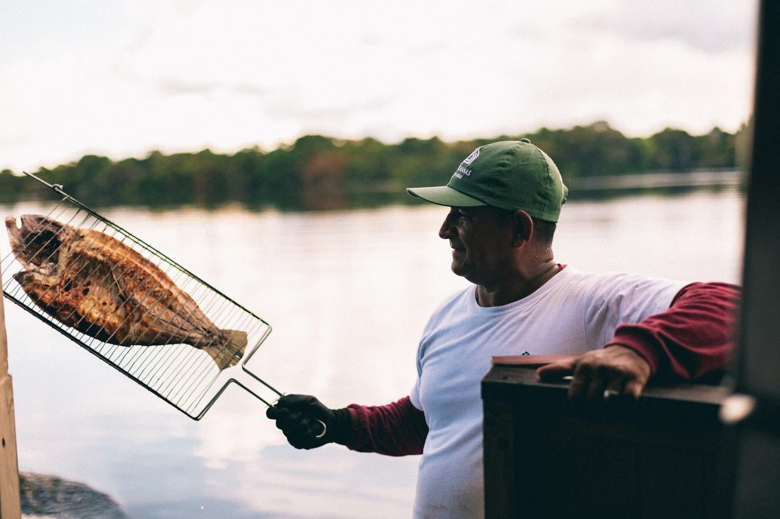 Peixe do rio, fogo de lenha e um sorriso. Assim é a cozinha ribeirinha de verdade. Um homem sorridente com boné verde segura um peixe inteiro grelhado em uma grelha metálica sobre o rio, com a floresta amazônica densa refletida na água calma ao fundo