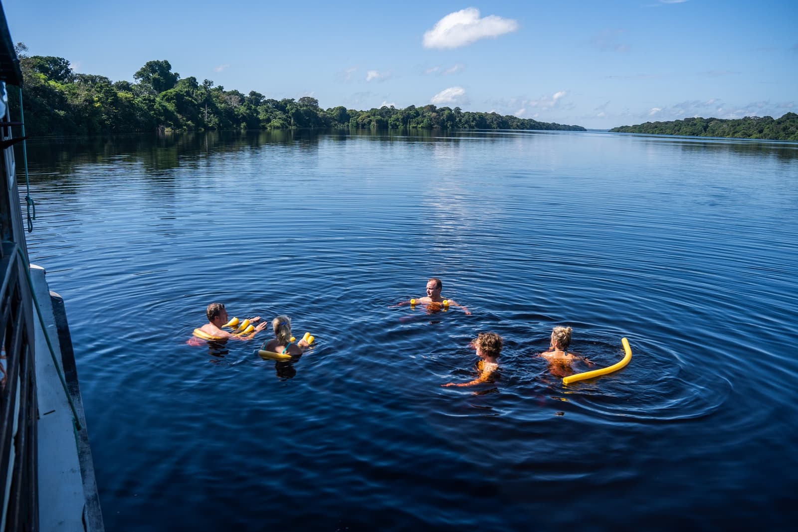 Mergulhar nas águas pretas da Amazônia é uma dessas coisas que a gente não esquece de fazer. Cinco hóspedes nadando nas águas escuras do Rio Negro ao lado de um barco tradicional na Anavilhanas Jungle Lodge Brazil, com macarrões flutuadores amarelos e floresta amazônica densa às margens sob céu azul