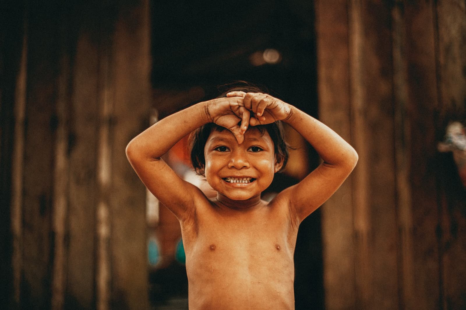 Tem sorriso que aparece antes mesmo de você pedir. Uma criança sorridente com as mãos erguidas de forma brincalhona diante de uma parede de madeira em uma comunidade ribeirinha próxima ao Anavilhanas Jungle Lodge Brasil
