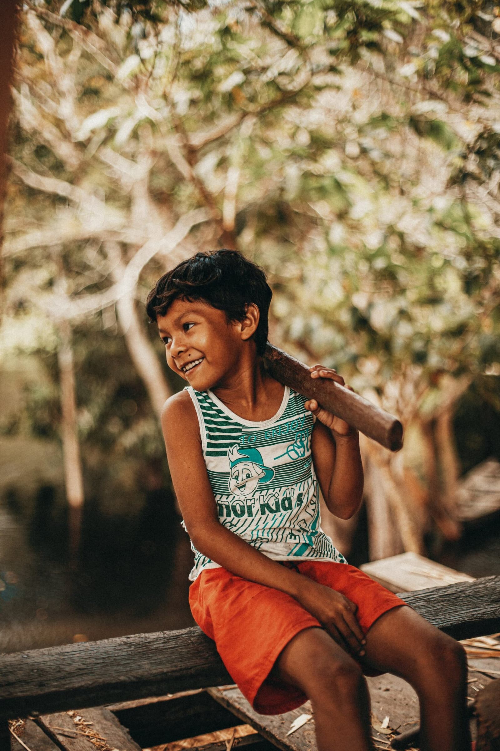 A tarde rendeu, e ele sabe disso melhor do que ninguém. Um menino sorridente senta em uma viga de madeira segurando um galho no ombro durante a visita a uma comunidade ribeirinha perto do Anavilhanas Jungle Lodge Brazil, com árvores ao fundo banhadas de sol