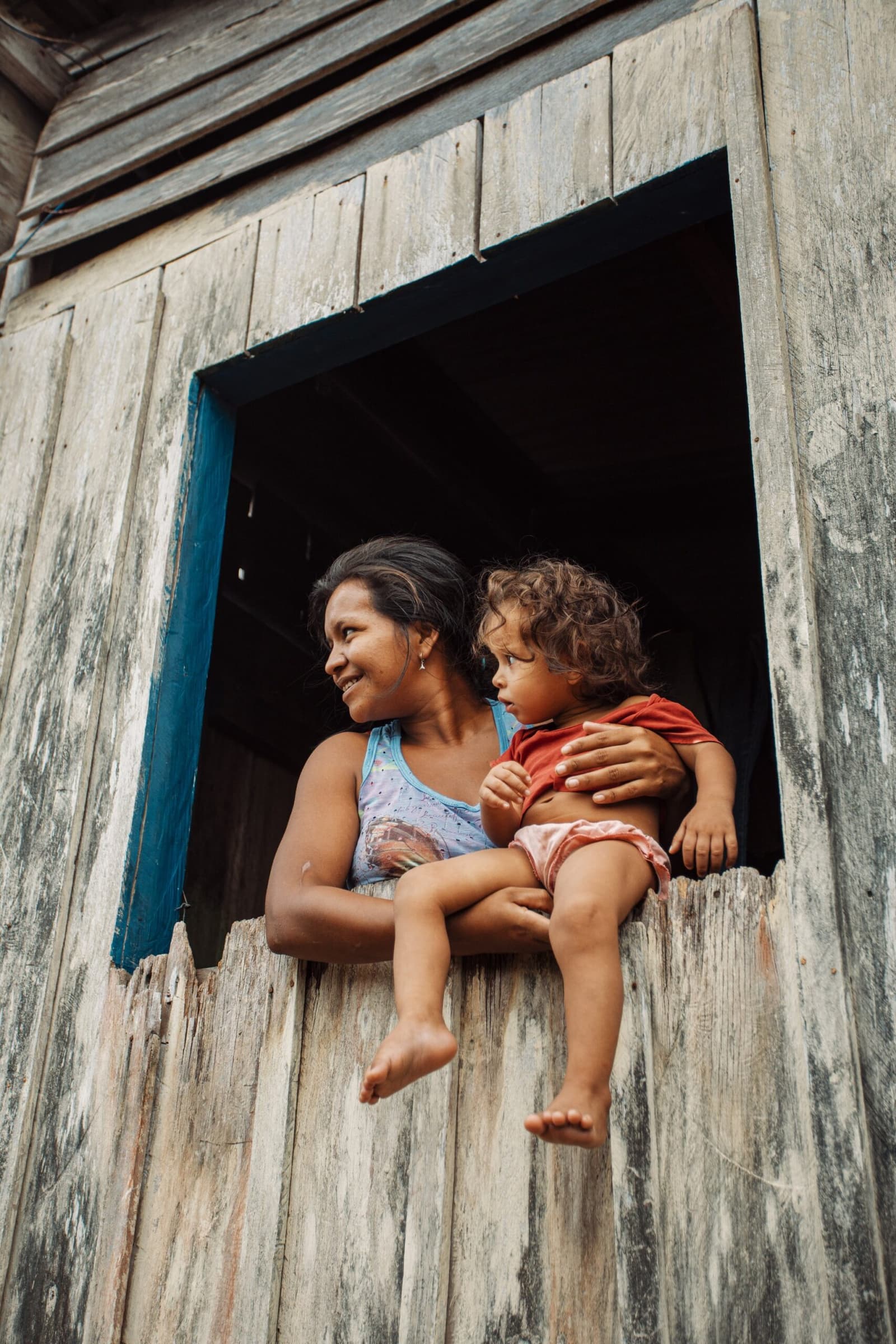 A Amazônia que importa mora nos sorrisos de quem vive nela. Uma mulher sorridente segura uma criança descalça no parapeito de uma janela de madeira envelhecida durante uma visita a uma comunidade ribeirinha próxima à Anavilhanas Jungle Lodge Brasil
