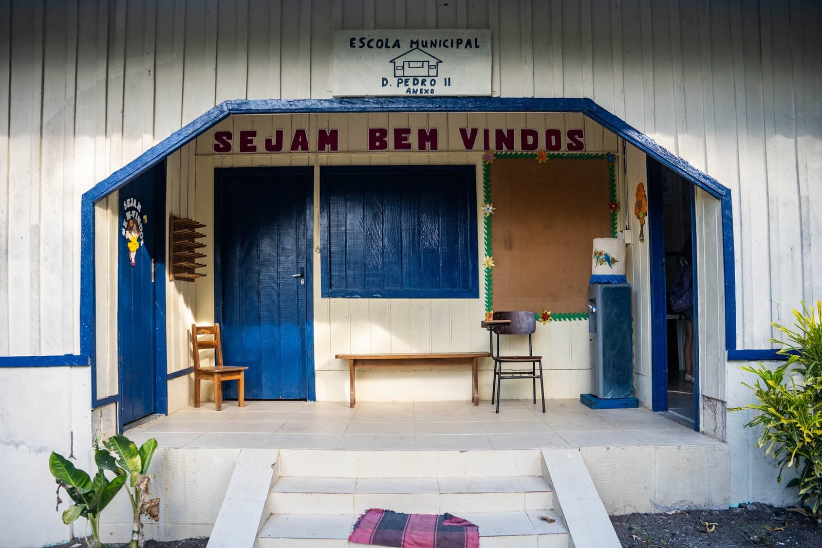 Escola pequena, recado claro: todo mundo é bem-vindo aqui. Entrada da Escola Municipal D. Pedro II, uma escola de madeira branca com detalhes azuis e a faixa 'Sejam Bem Vindos' em uma comunidade ribeirinha da Amazônia