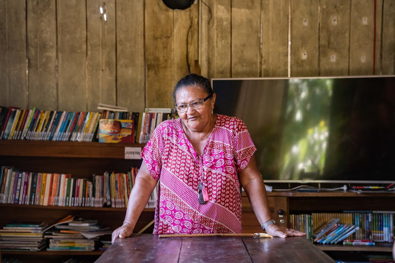 Ela sabe o nome de cada livro naquelas prateleiras, e de cada criança que os pegou emprestado. Uma moradora local com vestido rosa estampado apoia-se numa mesa de madeira dentro de uma biblioteca comunitária próxima à Anavilhanas Jungle Lodge Brazil, com estantes coloridas nas paredes de madeira ao fundo