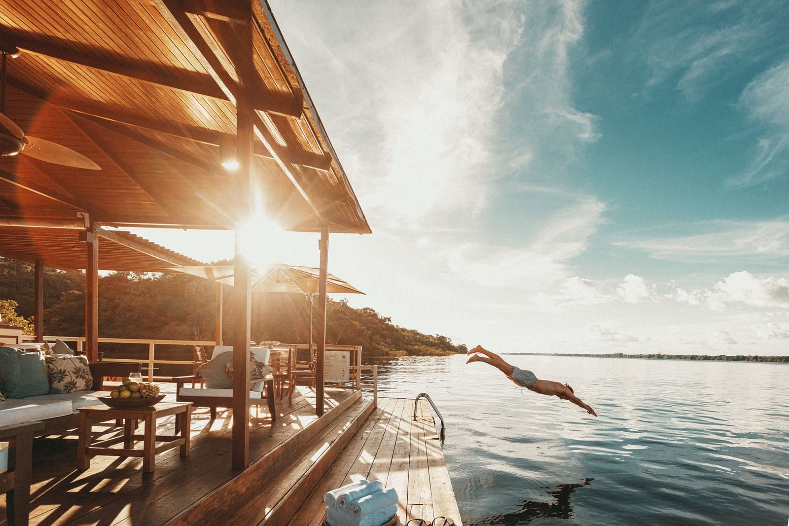O rio está ali. A decisão se toma sozinha. Um hóspede mergulha do deck de madeira da Anavilhanas Jungle Lodge Brazil nas águas calmas do Rio Negro na hora dourada, com uma área de estar ao ar livre e floresta exuberante ao fundo