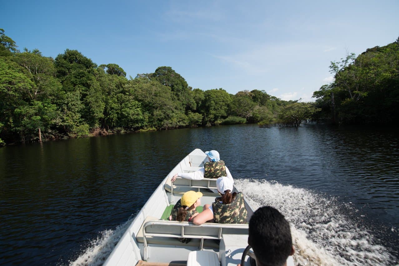 A floresta de cada lado, e a próxima curva como único destino. Visitantes com coletes salva-vidas navegam em alta velocidade por um rio de águas negras da Amazônia em uma excursão de barco a motor pelo Anavilhanas Jungle Lodge Brazil, ladeados por densa floresta tropical sob céu azul
