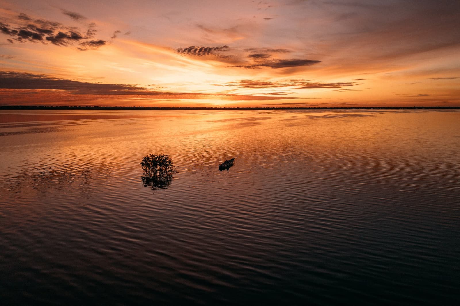 Uma canoa, uma árvore encharcada e o rio que resolve virar espelho. Um pequeno barco a motor desliza sobre as águas calmas do Rio Negro ao amanhecer durante um passeio de nascer do sol na Anavilhanas Jungle Lodge Brasil, com um arbusto submerso em silhueta sob um céu alaranjado e carmesim