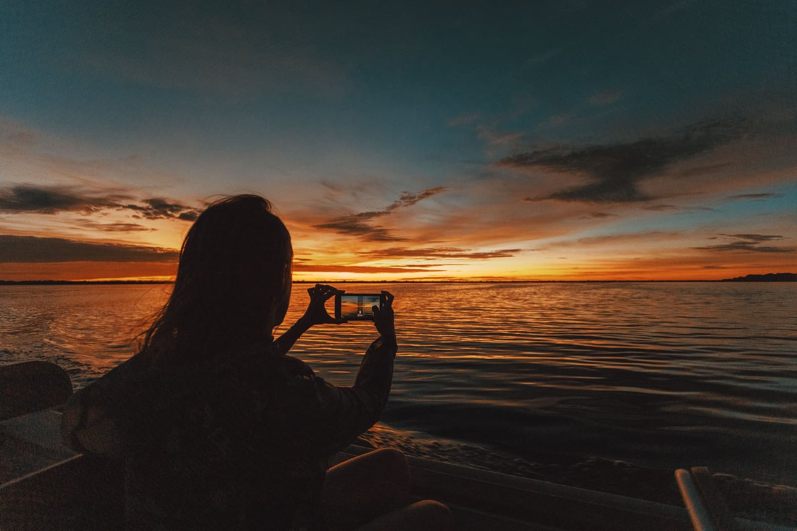Tem pôr do sol que a gente sabe que o olho não vai guardar sozinho. Silhueta de uma viajante fotografando um pôr do sol alaranjado e intenso sobre o rio largo a partir de um barco na Anavilhanas Jungle Lodge Brasil
