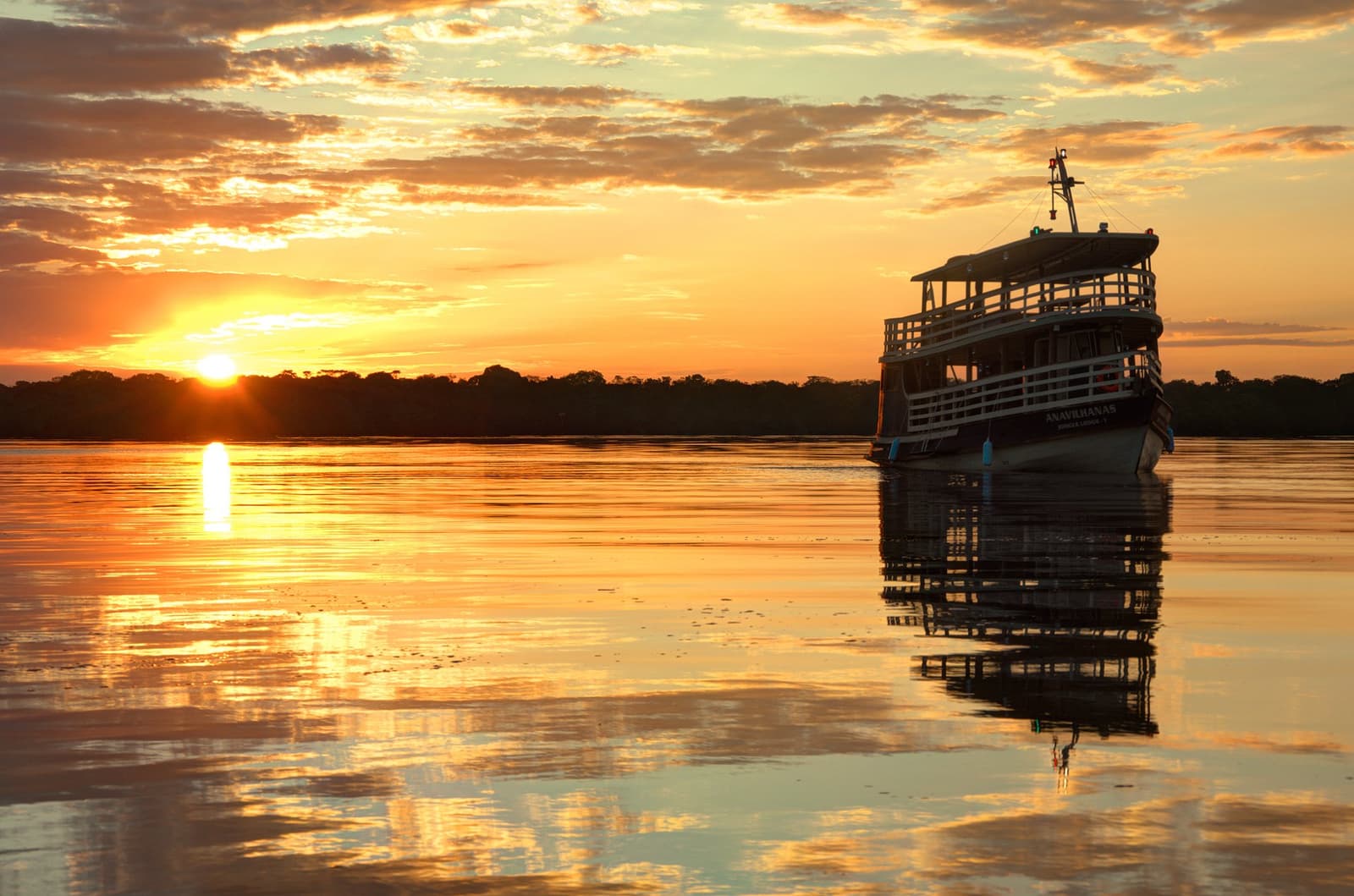 Rio parado, céu em chamas, e a viagem ainda mal começou. O barco de expedição da Anavilhanas Jungle Lodge Brazil ancorado no Rio Negro espelhado sob um pôr do sol alaranjado e flamejante, com reflexo perfeito na água calma