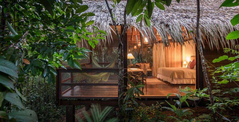 A warmly lit thatched-roof bungalow at Anavilhanas Jungle Lodge Brazil, with a woven hammock on the open deck and a king bed visible through floor-to-ceiling glass walls, surrounded by dense rainforest foliage at dusk A warmly lit thatched-roof bungalow at Anavilhanas Jungle Lodge Brazil, with a woven hammock on the open deck and a king bed visible through floor-to-ceiling glass walls, surrounded by dense rainforest foliage at dusk