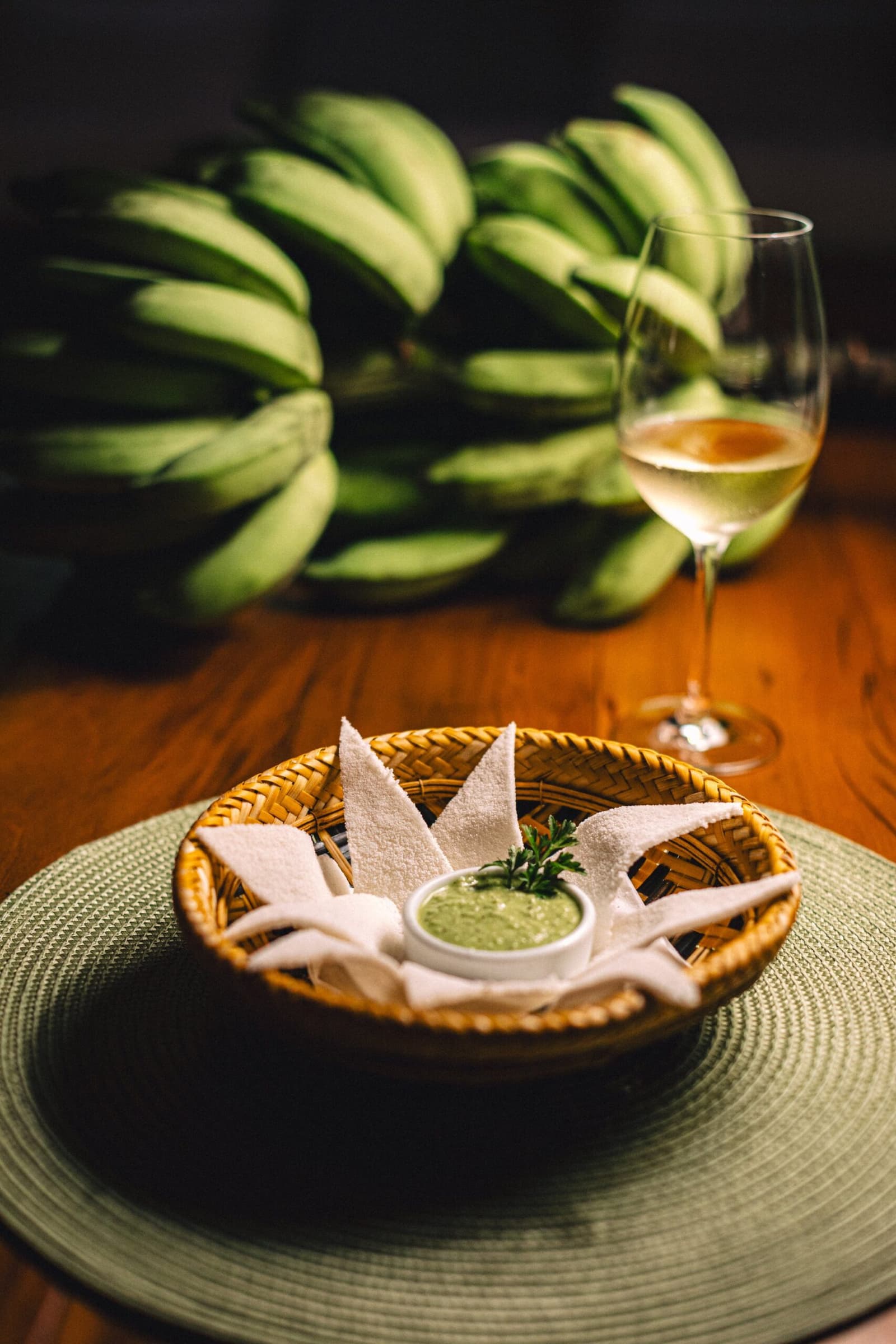 Chegou, se acomode: o bem-vindo da Amazônia começa na mesa. Uma cesta de rattan trançado com pedaços de pão branco dispostos ao redor de um pequeno recipiente de molho verde de ervas, servido na Anavilhanas Jungle Lodge Brazil ao lado de uma taça de vinho branco, com um cacho de bananas verdes desfocado ao fundo