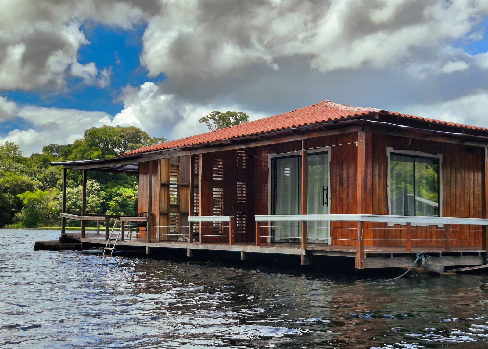 Flutuando sobre o rio escuro, com a floresta logo ali — bem-vindo à sua casa na Amazônia. Uma pousada flutuante de madeira sobre água escura de rio, com deque envolvente, venezianas e portas de vidro deslizantes, rodeada pela mata fechada — hospedagem em um tour pela selva amazônica brasileira