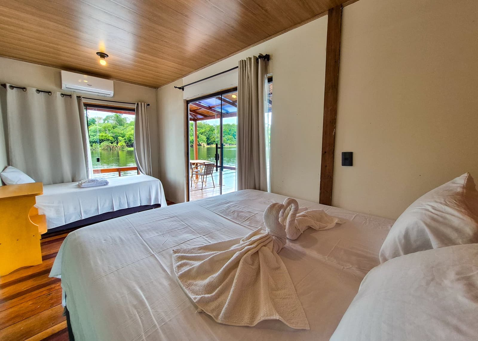 Cisnes de toalha, rio quieto e Amazônia a passos de distância. Quarto duplo em lodge flutuante durante um tour pela selva amazônica brasileira, com roupa de cama branca, decoração de toalha em forma de cisne na cama de casal, teto de madeira, ar-condicionado e porta de vidro deslizante abrindo para varanda com vista para o rio e a floresta