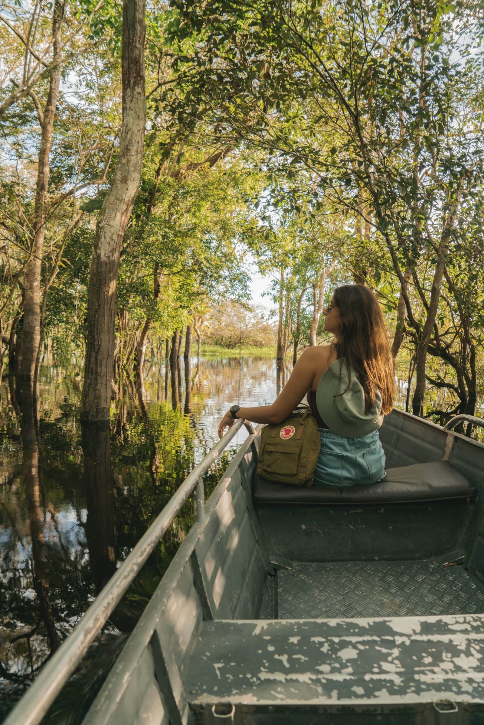 A floresta alagada engole o barco em silêncio — só luz, água e espanto. Photo: Yuri Panichi Uma viajante sentada na proa de um pequeno barco de metal durante um passeio pela selva amazônica brasileira, contemplando uma floresta inundada com árvores refletidas na água escura e parada