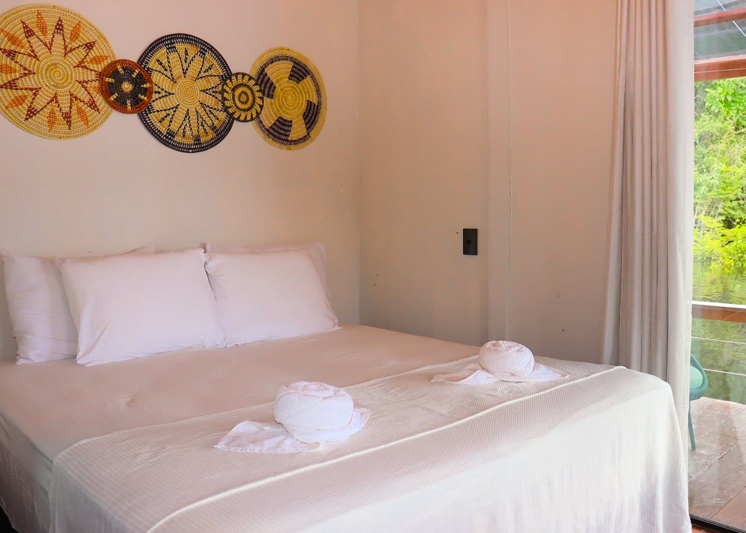 Artesanato local nas paredes, lençóis frescos e a floresta a poucos metros. Uma cama de casal com roupa de cama branca e toalhas dobradas sob decoração de cestos trançados na parede de um quarto de lodge confortável, com janela voltada para a floresta verde