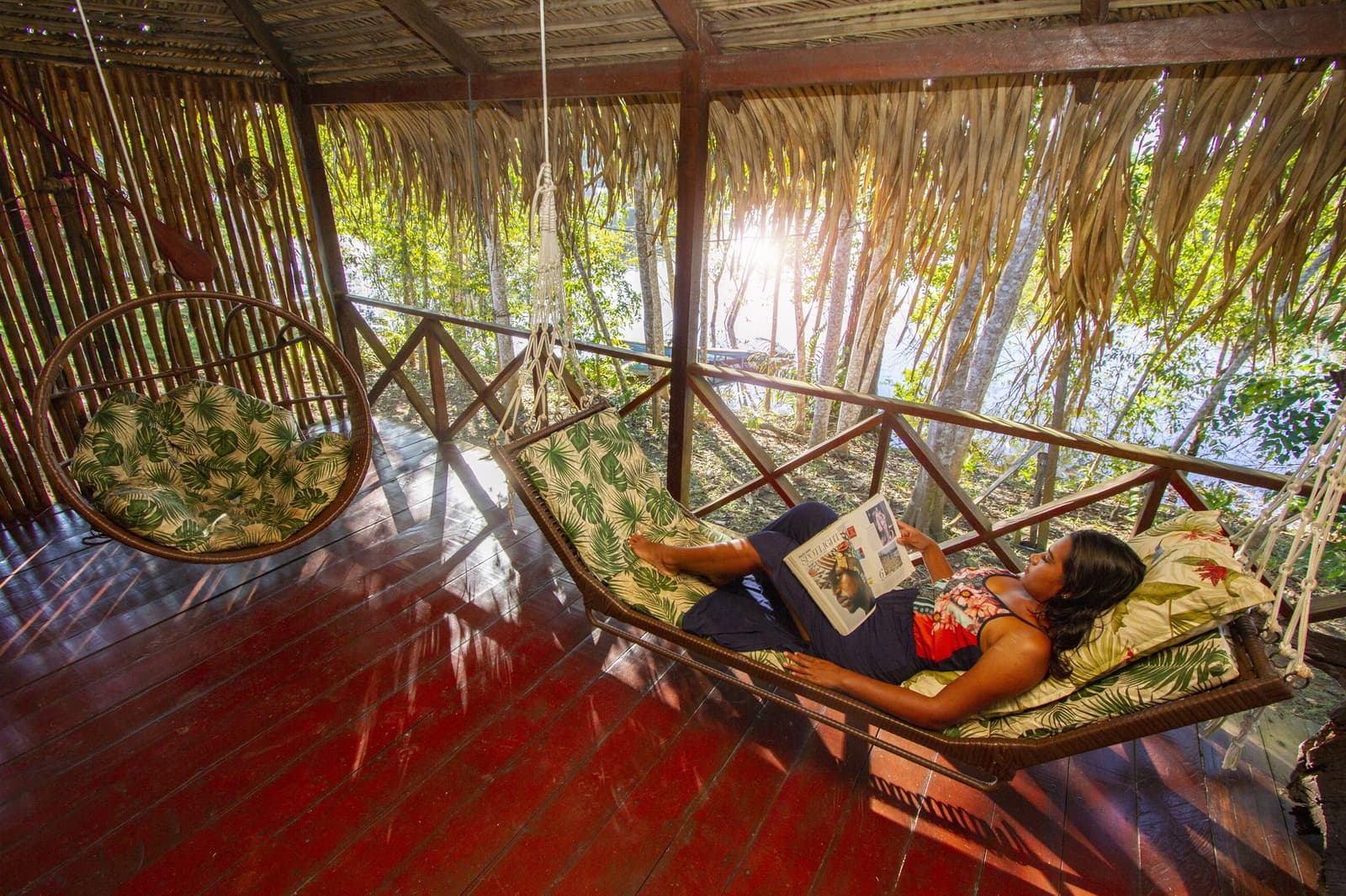 Balanço, brisa e mata fechada: o descanso perfeito na Amazônia. Photo: David Nascimento @ Uma hóspede deita em uma rede de rattan lendo uma revista em uma varanda coberta de palha, com folhagem tropical e luz do sol filtrando pelas árvores ao fundo