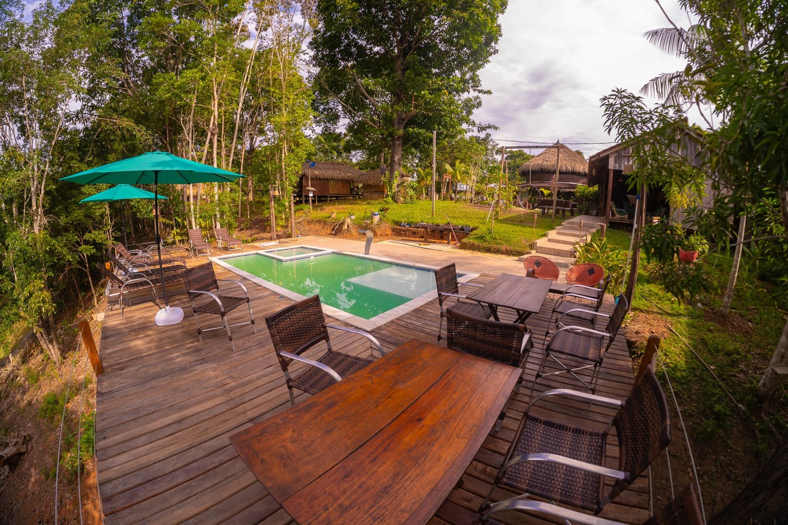 Mergulho rápido antes do próximo roteiro na floresta. Um deck de madeira envolve uma piscina retangular em um lodge de tour pela selva amazônica brasileira, com cadeiras de rattan, guarda-sol turquesa, espreguiçadeiras e bangalôs com telhado de palha entre árvores da floresta tropical