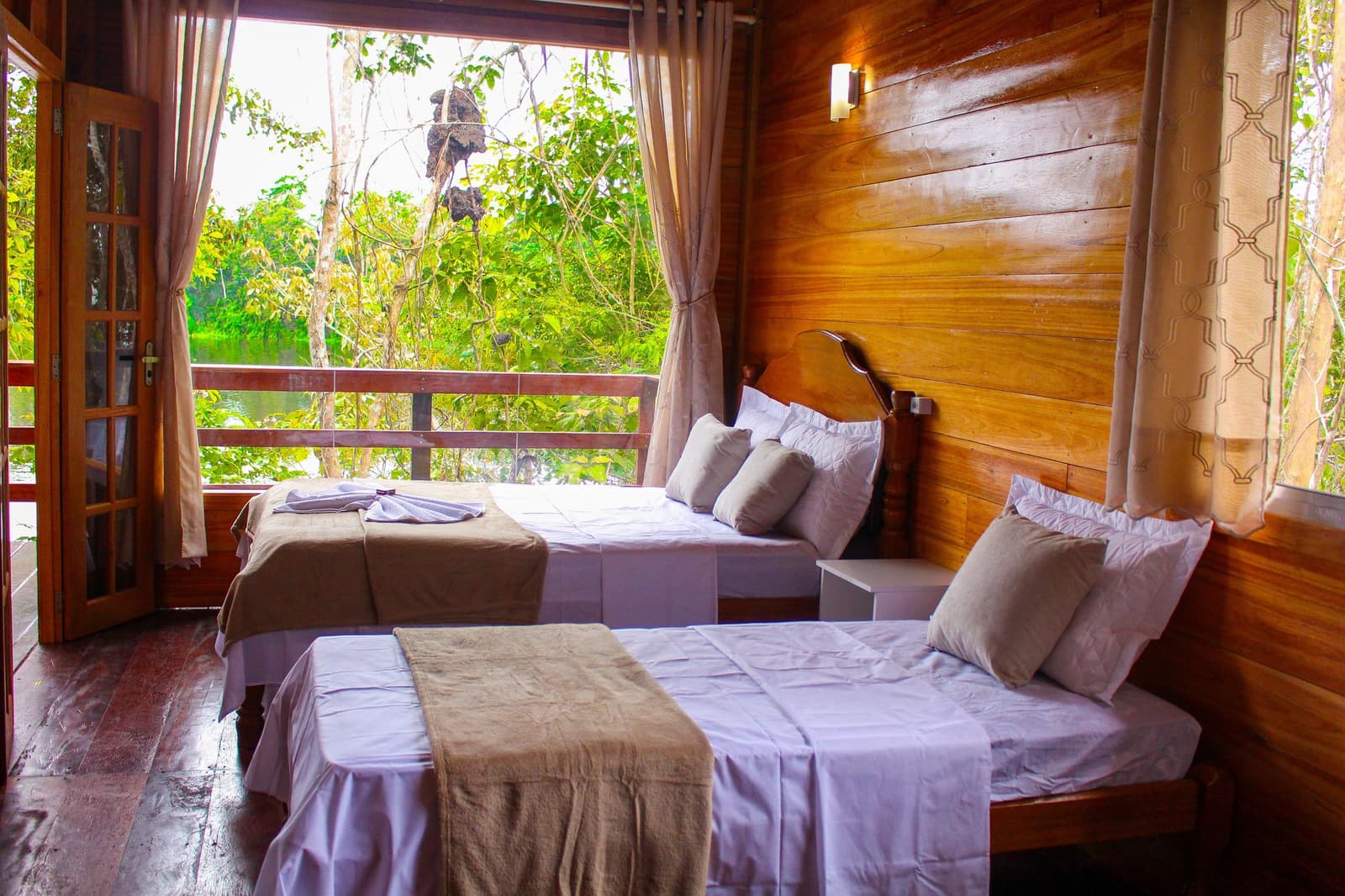 Cama arrumada, madeira aconchegante e a Amazônia logo além da varanda. Quarto duplo com roupas de cama brancas e paredes de madeira em um lodge na selva amazônica, com varanda abrindo para um igarapé verdejante cercado por árvores da floresta tropical
