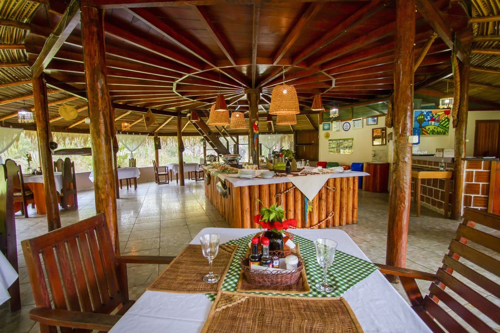 Refeições feitas com capricho no coração da floresta amazônica. Pavilhão de jantar ao ar livre com teto de palha em lodge na selva amazônica, com colunas rústicas de madeira, luminárias de vime, mesas com toalhas brancas e balcão de buffet com frutas tropicais